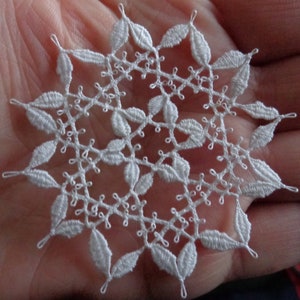 May include: Delicate white lace snowflake with a detailed floral design. The snowflake is made of intricately woven threads, featuring leaf-shaped elements and a circular pattern. The lace is held in a hand, showcasing the fine craftsmanship.