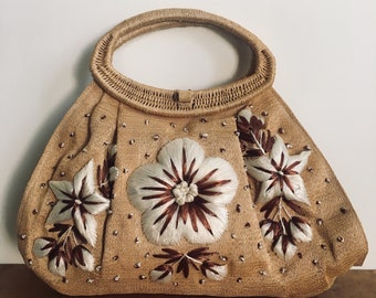 Könnte beinhalten: Eine braune Strohtasche mit weißen und braunen gestickten Blumen. Die Tasche hat einen oberen Henkel und eine abgerundete Form.