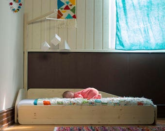 May include: A newborn baby in a red and white striped outfit lies on a wooden crib with a colorful quilt. The crib is in a room with a wooden wall and a hanging mobile with geometric shapes.