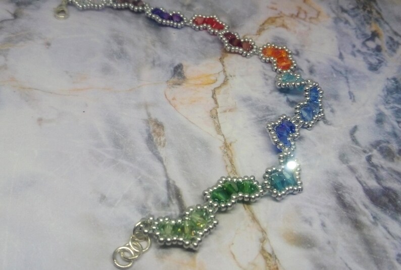 May include: A rainbow-coloured beaded bracelet with silver beads and a silver clasp. The bracelet is made of small, faceted beads that are arranged in a zig-zag pattern.