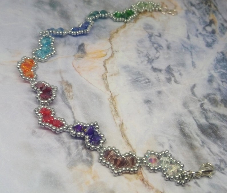 May include: A rainbow-coloured beaded bracelet with a silver clasp. The beads are arranged in a heart shape and are connected by silver wire.