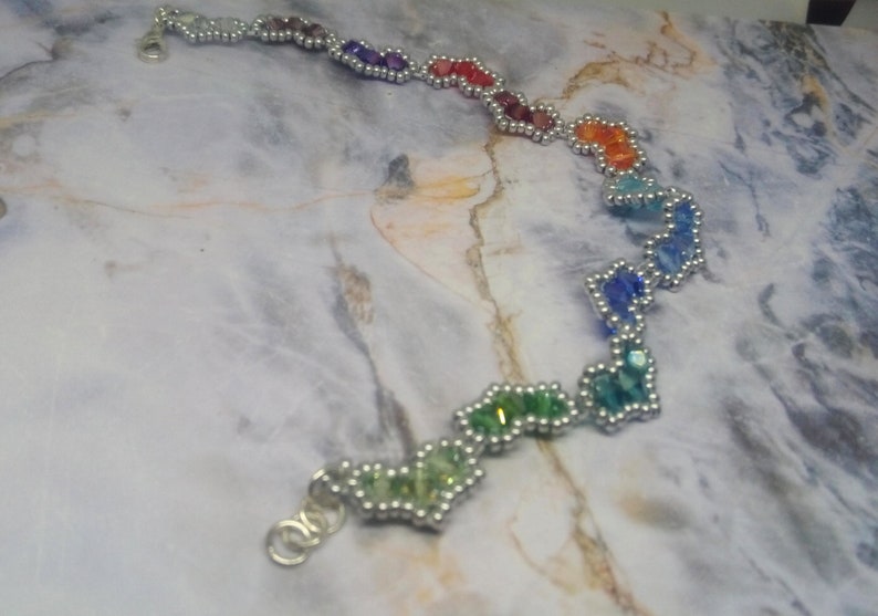 May include: A rainbow-coloured beaded bracelet with silver accents. The beads are arranged in a heart shape and are connected by silver wire.