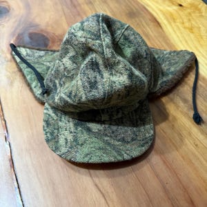 May include: A camouflage-patterned hat with ear flaps and a brim. The hat is primarily green and brown, with a textured fabric. Black drawstrings are attached to the ear flaps.