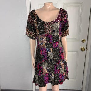 May include: A patchwork mini dress with a scoop neckline and short sleeves. The dress features a mix of floral and paisley patterns in shades of black, purple, pink, and cream. The dress has a ruffled hem and is displayed on a mannequin.