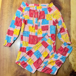 May include: A colorful patchwork pajama set with a long-sleeved top and matching pants. The set features a mix of red, yellow, and blue squares with white stripes. The top has a button-down front and the sleeves have ruffled cuffs.