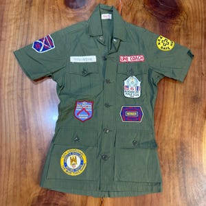 May include: Olive green short-sleeved shirt with multiple patches. Patches include "LINE COACH", "MEMBER", and "BANGICK JR. RIFLE CLUB". The shirt has a name tag that reads "ROBINSON".