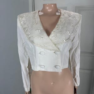 May include: A white, cropped blazer with a decorative, embroidered collar. The blazer features a double-breasted design with four white buttons. The collar has a scalloped edge and intricate floral embroidery.