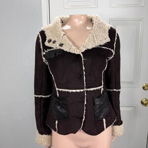 May include: A dark brown jacket featuring a cream-colored faux shearling collar and cuffs. The garment has exposed white seams, a button-up front, and two black textured pockets on the lower front, along with a smaller pocket on the upper right chest.