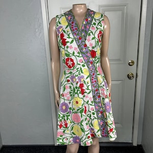 May include: A sleeveless knee-length dress featuring a white base with a vibrant floral pattern in red, yellow, pink, and purple. The dress has a V-neckline and a distinct diagonal band of purple floral trim that runs from the shoulder to the hem.