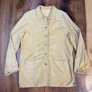 May include: A tan suede jacket with a collared neckline, long sleeves, and two front pockets. The jacket has a button-up closure and buttons on the cuffs. The garment is laid flat on a wooden surface.