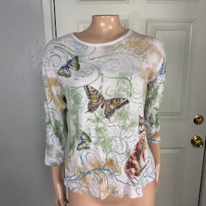 May include: White long-sleeve shirt with a round neck, featuring butterfly and floral designs in shades of green, yellow, and blue. The shirt has a relaxed fit and three-quarter sleeves. The design includes a cat illustration.