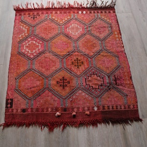May include: A red and orange Moroccan rug with a geometric pattern of hexagons and crosses. The rug has a fringe border.
