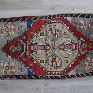 May include: A small, rectangular rug with a traditional Persian design. The rug is primarily red with a central medallion in beige, green, and red. The rug has a fringe border.
