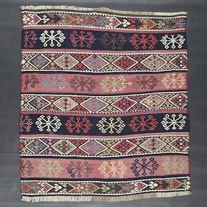 Puede incluir: Una alfombra rectangular tejida a mano con un patrón geométrico. La alfombra presenta bandas alternas de azul marino y rosa empolvado, adornadas con motivos de diamantes y copos de nieve en varios colores, incluyendo rojo, blanco y verde. Los bordes tienen flecos.