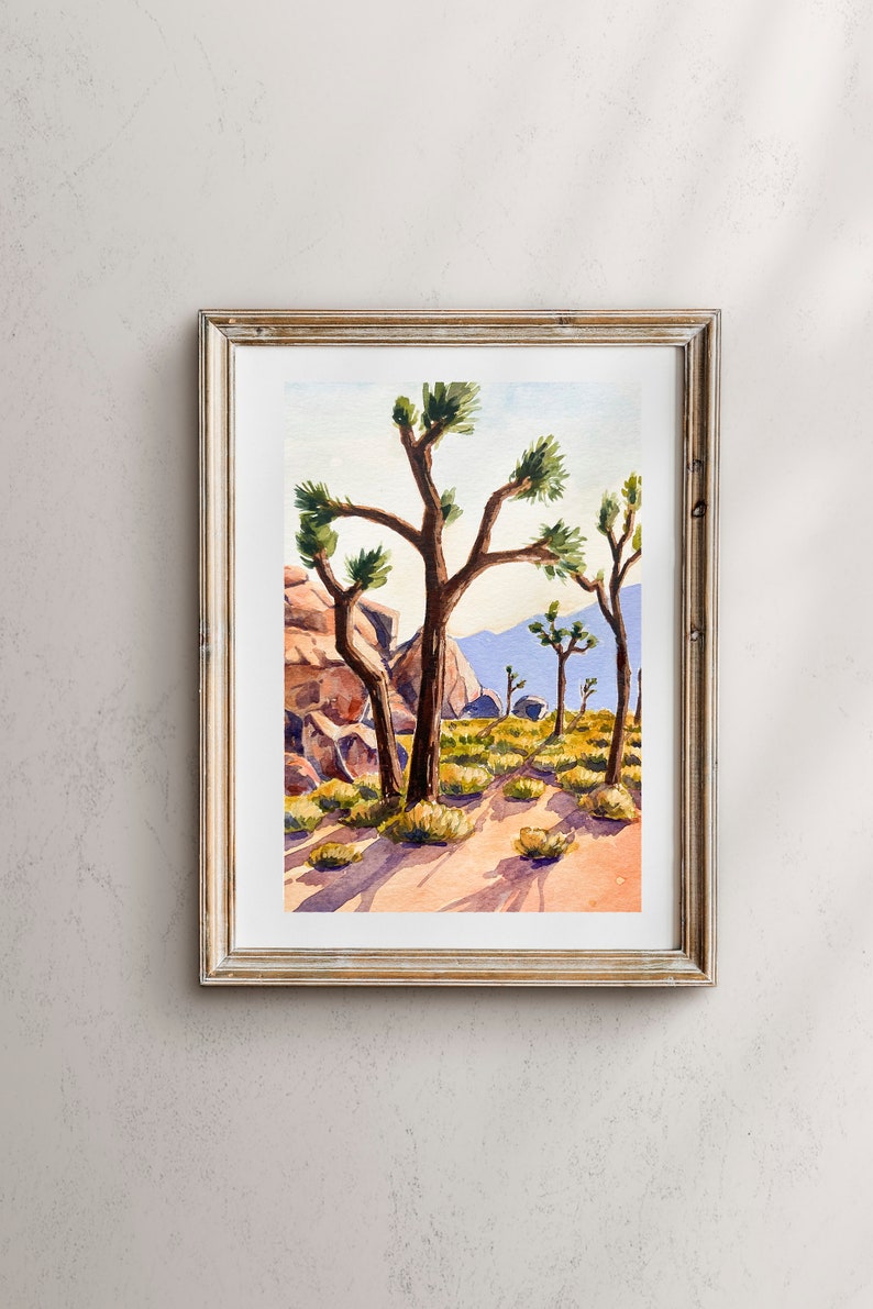 May include: Watercolor painting of a desert landscape with Joshua trees. The artwork is framed in a gold-colored frame and features a path, rocks, and mountains in the background. The painting is in a vertical orientation.