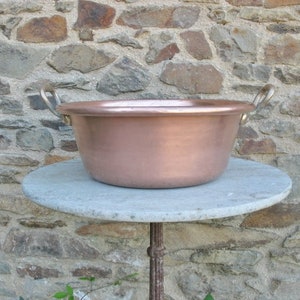 May include: A large copper bowl with two handles sits on a gray stone table. The bowl is round and has a smooth, shiny surface. The handles are made of metal and are attached to the sides of the bowl.