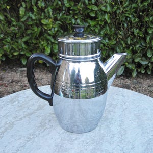 May include: A silver chrome coffee pot with a black handle and lid. The pot is sitting on a gray table.