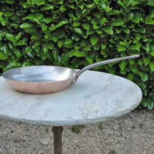 May include: A copper frying pan with a long handle sits on a white marble table. The pan is slightly dented and has a worn, aged look.