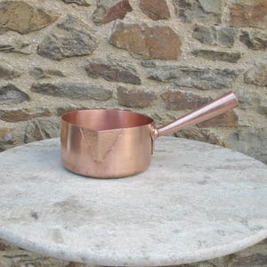 May include: A polished copper saucepan with a single long handle and a V-shaped pouring spout. The pan features a smooth, metallic finish and a cylindrical body, showcasing the warm, reddish-brown tones of the copper material throughout.