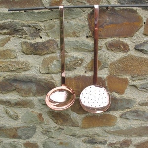 Puede incluir: Dos utensilios de cocina de cobre colgados de un estante de metal contra una pared de piedra. El utensilio de la izquierda es una cuchara sólida con un cuenco redondeado. El utensilio de la derecha es una cuchara ranurada con un cuenco redondeado.