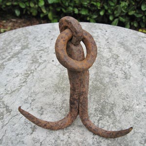 May include: A rusty metal hook with a double loop at the top. The hook is resting on a gray concrete surface.