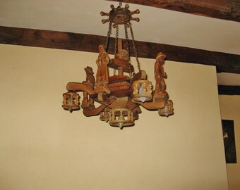 May include: A wooden chandelier with intricate carvings and figures, suspended from chains. The light fixture features multiple arms with small lamps and is made of light brown wood. The chandelier is hanging from a ceiling with wooden beams.