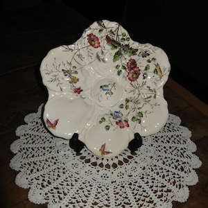 May include: A white ceramic oyster plate with a floral design. The plate has six sections, each with a different floral motif. The plate is decorated with butterflies and flowers in shades of pink, red, yellow, and blue.