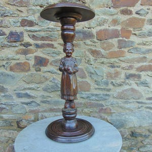 May include: A dark brown wooden pedestal stand with a carved figure of a woman holding a basket. The stand has a round top and a base with a decorative design.