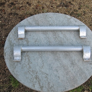 May include: A round, gray metal lid with two parallel, silver handles. The lid has a textured surface and is likely used for covering a manhole or similar access point. The handles are securely attached to the lid.