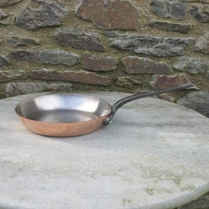 Op de afbeelding: Een koekenpan met koperen bodem, een roestvrijstalen binnenkant en een lange, zwarte handgreep. De pan staat op een ronde, lichtgekleurde stenen tafel. De achtergrond is een stenen muur.