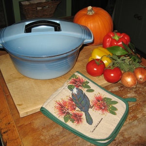 Op de afbeelding: Een blauwe geëmailleerde gietijzeren pot met een deksel op een houten snijplank. Er liggen groenten op de plank, waaronder tomaten, uien en paprika's. Een pompoen en een groen-witte ovenwant met een vogelontwerp zijn ook op de afbeelding te zien.
