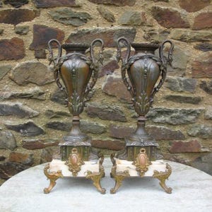 May include: Two antique bronze urns with ornate handles and decorative detailing. The urns sit atop white marble bases with gold-colored feet. The background is a stone wall, and the urns are displayed on a round table.