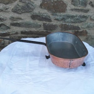 Puede incluir: Una sartén antigua de cobre con un mango largo y negro y forma rectangular. El interior de la sartén es plateado y se apoya sobre pequeños pies negros. El exterior de cobre muestra signos de edad y uso. La sartén se exhibe sobre un paño blanco.