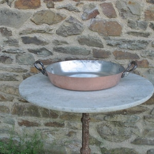 May include: A copper pan with two black handles sits on a round, light gray stone table. The pan is empty and has a shiny, reflective surface.