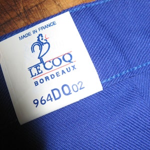 May include: A white tag with blue and red text that reads "Made in France", "Le Coq", "Bordeaux", and "964DQ02". The tag is attached to a blue fabric.