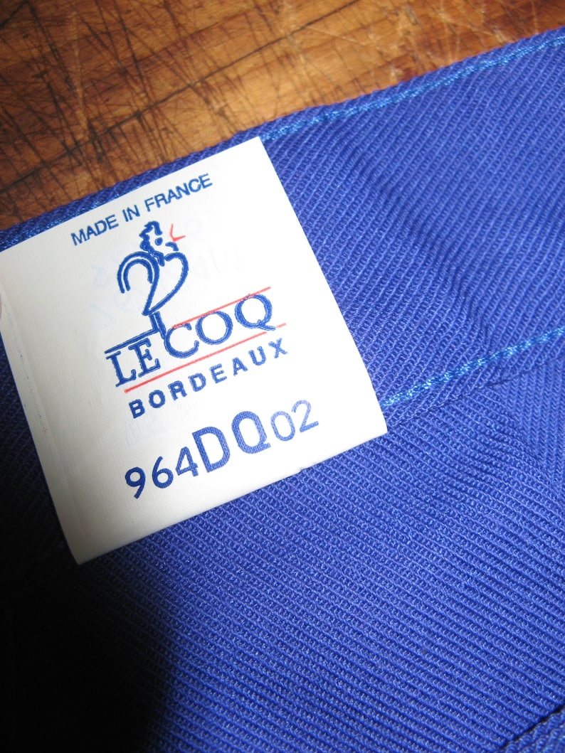 May include: A white tag with blue and red text that reads "Made in France", "Le Coq", "Bordeaux", and "964DQ02". The tag is attached to a blue fabric.