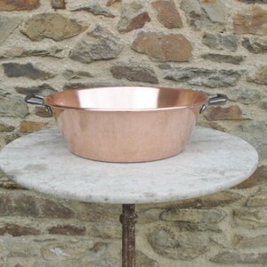 May include: A large copper bowl with two handles sits on a round, light gray stone table. The bowl is shiny and has a slightly hammered texture. The table is on a stone wall background.