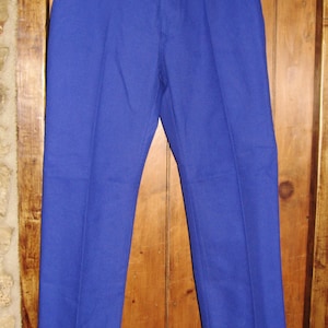 May include: A pair of blue work pants hanging on a wooden hanger. The pants have a button fly and are made of a thick, durable fabric.