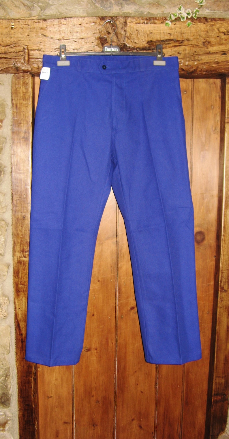 May include: A pair of blue work pants hanging on a wooden hanger. The pants have a button fly and are made of a thick, durable fabric.