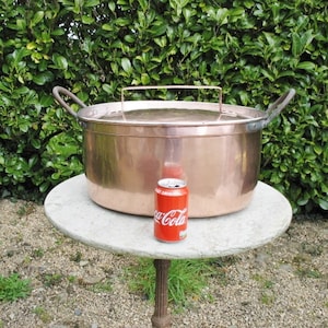 May include: Large copper pot with a lid and two handles. The pot is sitting on a white marble table top. A can of Coca-Cola is next to the pot for scale.