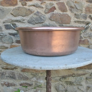 May include: A large copper bowl with two brass handles sits on a round, light gray stone table. The bowl is empty and has a shiny, reflective surface.