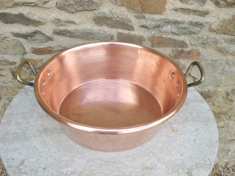 May include: A large copper pot with two handles. The pot is sitting on a light gray stone surface.