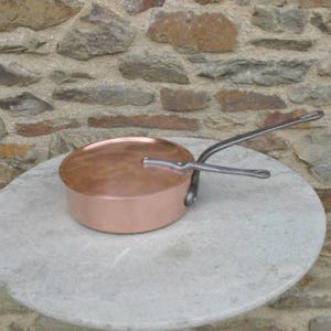 Pode incluir: Uma panela de cobre com uma tampa e uma alça longa e curva. A panela está sobre uma superfície cinza claro.