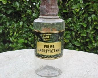 May include: A clear glass bottle with a rusty metal lid and a label that reads "Pulvis Anth:Pyrethri". The bottle is sitting on a light gray surface.