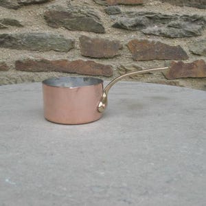 May include: A small copper saucepan with a brass handle. The saucepan is sitting on a gray stone surface.