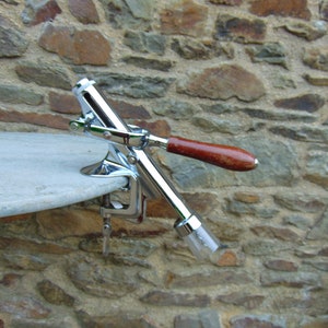 May include: A silver and brown metal bottle opener with a lever and a clamp. The opener is attached to a wooden surface.