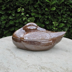 May include: A brown ceramic serving dish shaped like a duck. The dish is resting on a light grey stone surface.