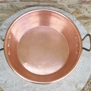 May include: A large, round copper pan with two handles. The pan is shiny and has a smooth surface.