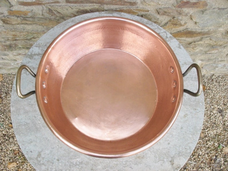 May include: A large, round copper pan with two handles. The pan is shiny and has a smooth surface.