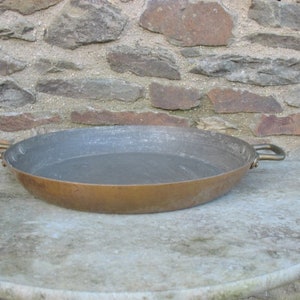 May include: A large, round copper pan with two handles. The pan is sitting on a light gray stone surface.